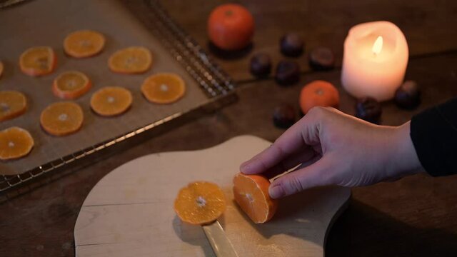 weibliche Hand schneidet Orange, Zitrusfrucht in Scheiben und legt sie zum trocknen auf ein Backpapier. weihnachtliche Stimmung und Vorbereitung für Weihnachten mit Kerzenlicht.