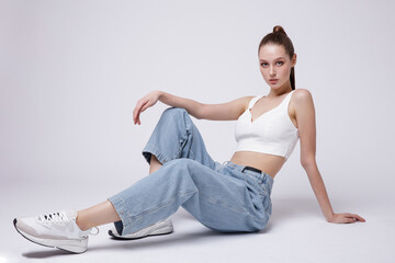 High fashion photo of a beautiful elegant young woman in a pretty white top and sneakers, blue denim jeans posing over white, soft gray background. Studio Shot. The model is sitting
