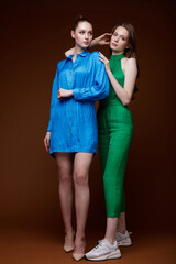 Two fashion models in short blue dress, shoes, green jumpsuit, sneakers. Beautiful young women. Studio shot, portrait. Brown background.