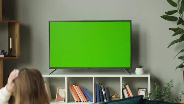 Woman watching television with chroma green screen. Unrecognizable female person switching channels on tv with remote controller, sitting on sofa in living room. Spare time at home, relaxing.