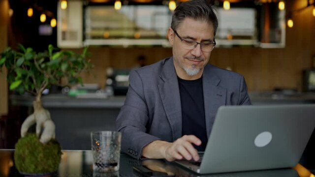 Businessman sitting at table in hotel lobby, restaurant or cafe, working on laptop computer. Mature age, middle age, mid adult man in business casual, confident happy smiling.