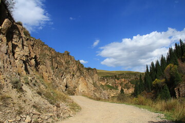 The road is high in the mountains on the Assy plateau, on the left is a sheer stone cliff with uneven edges, on the right is a mountain slope with fir trees, the sky is with clouds, summer, sunny