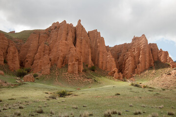 Red relief sandy-clay towers of a mountain ridge slope on the Assy high plateau, at the foot of the slope there is a green meadow with grass and bushes, a sky with gray clouds, summer