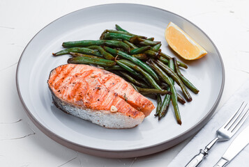 grilled salmon steak with string beans and lemon in a round white plate on a white background