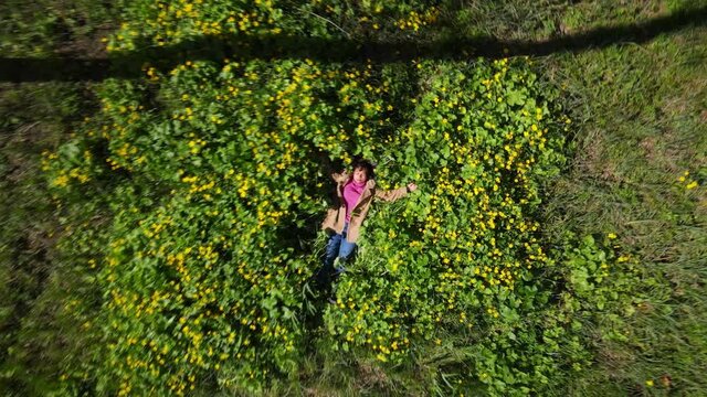 Ascending drone shot from a woman lying among a marsh-marigold flowerbed to the top overview of a temperate forest at spring, France