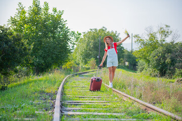 one woman in a hat and a suitcase walks along the sleepers