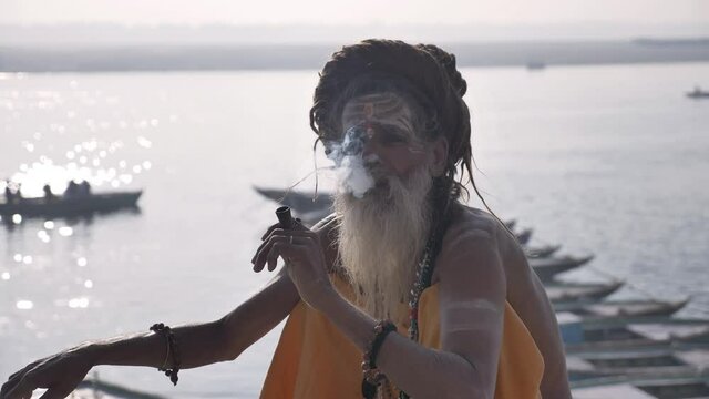 A bearded old or elderly sadhu sitting and smoking on the banks of the Ganga in Varanasi, India. 