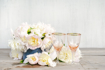 Beautiful celebration composition: Two beautiful empty wineglass, bouquet of white peonies on rustic wooden background; copy space