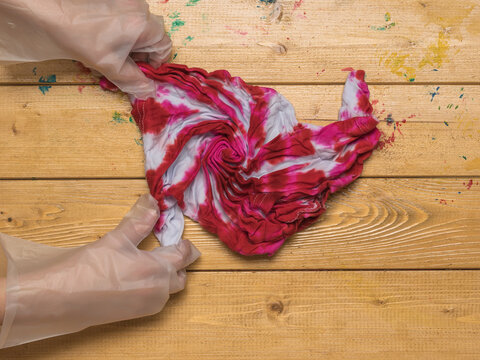 A girl in gloves unfolds a T-shirt painted in tie dye style. Flat lay.