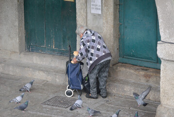 Pessoa pobre e idosa a alimentar pombos numa zona velha de cidade, puxa um carro de mão com milho...