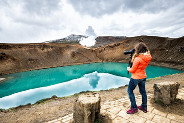 Woman photographer camera on Viti Crater of Krafla caldera geothermal formation with small lake of blue turquoise green water and mountains snow during summer