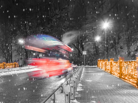 A red train rides across the bridge. Garlands burn beautifully on the fence and it snows. Part of the photo is black and white, part is color. Christmas atmosphere. Paravoz blurred traffic. Soft focus