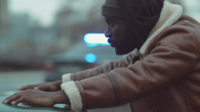 Upset African American man standing outdoors by car and waiting for police search police car with lights flashing in background