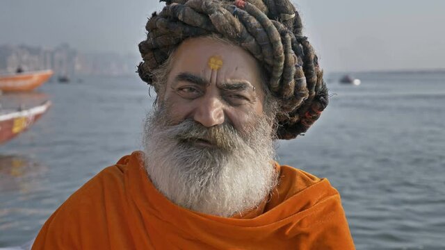 A bearded old or elderly male sadhu or Hindu priest wearing traditional clothes is sitting next to the Ganges River and looking at the camera while chanting a mantra or prayer in Varanasi