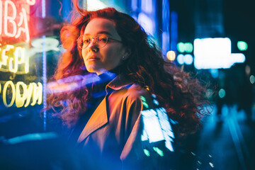 Curly haired young woman tourist with light makeup in glasses looks around standing near bar with colorful neon sign against night megalopolis - Powered by Adobe