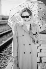 A young woman in a raincoat and dark glasses, with an umbrella in her hands. Retro style portrait at the railway station