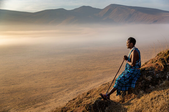 Masai Wwarrior sitting at the edge of one of Ngorongoro craters looking at the horizon and enjoying the surnrize
