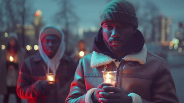 Selective focus shot of African American man holding candle in cup and looking at camera while standing outdoors with friend during candlelight vigil