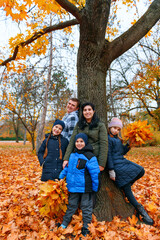 Portrait of a family with children in an autumn city park - happy people posing together near big tree, beautiful nature with yellow leaves as background.