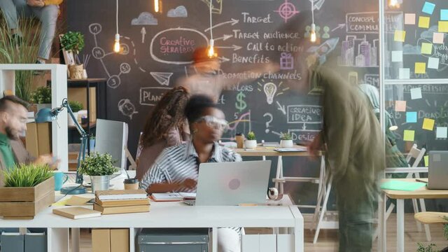 Zoom-in time lapse of office workers busy in creative workplace with chalkboard wall. Employees are talking using computers writing on board