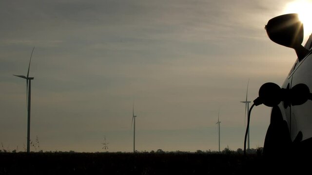 Windmills generate clean power in field. Sunlight shines over ev charging by wind energy at rural station against operating turbines close view