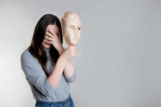 Complexes due to appearance or self-identification problems, concept. A young woman holds a mask in her hands hiding her face. Photos in light gray tones, gray background