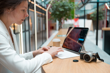 A photographer or retoucher works remotely with a laptop in a public space or coworking. A young brunette in a white jacket works with images, a designer or a social media manager