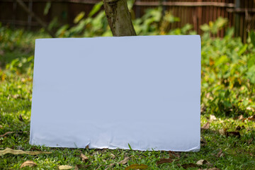 A white signboard leaning against a tree on the ground and the background behind it is blurred green.