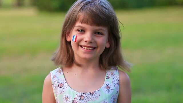 Adorable little girl smile at camera with cheeks painted in flag of France. Bastille day on July 14. The day of France. French summer holidays, travel concept.