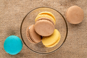 Several sweet macaroons in a glass plate on a burlap, close-up, top view.