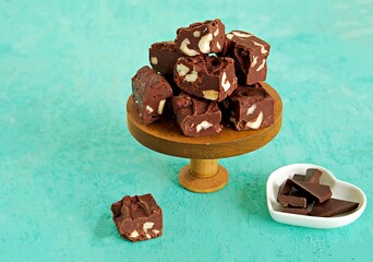 Sliced homemade fudge chocolates made from dark chocolate, condensed milk and hazelnuts on a wooden stand on a turquoise concrete background. Candy recipes. American cuisine.