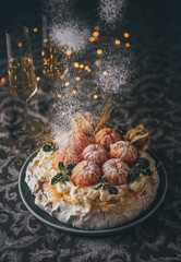 Pavlova cake low light still life pouring with powder sugar meringue-based dessert named after the Russian ballerina decorated with fresh mandarins, caramel, mint, physalis with Christmas lights