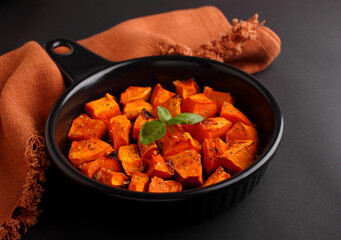 Pumpkin cut into pieces baked in a black round frying pan with a handle on a black background.