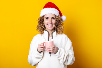 Smiling curly young woman in a santa hat with a cup on a yellow background.