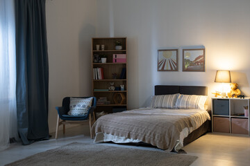 Empty bedroom with electric lamp on shelves, cozy armchair, pictures above bed at night