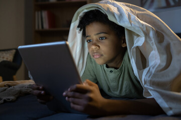 Concentrated African American boy with duvet on head lying in bed and using tablet while reading...