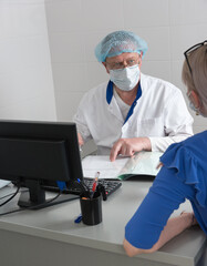 A doctor in white coat, mask and cap is sitting at a table in the office talking to a patient