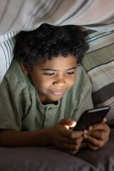 Content African American boy with curly hair addicted to gadget watching video on smartphone under duvet