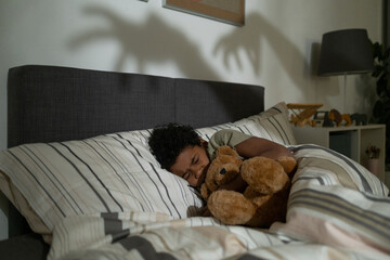 Scared African American boy embracing bear toy while sleeping in bed and having nightmares