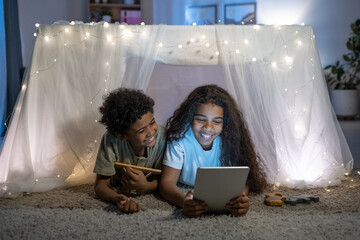 Cheerful African American brother and sister lying on carpet behind table covered with fabric and lights and using tablet