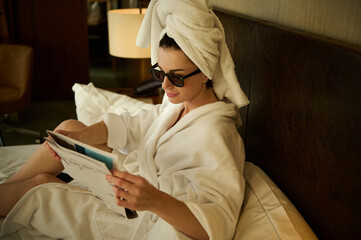 Overhead view of a Caucasian pretty woman wrapped in white bath towel reading news on newspaper and resting, after relaxing bathing, on the bed in the hotel bedchamber