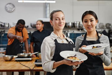 Waist up portrait of two young women wearing aprons and holding gourmet dishes in professional kitchen, copy space