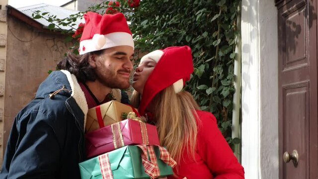 Young man in Santa Claus hat brings gifts to his girlfriend. She smiles at her boyfriend and kisses him on the cheek.