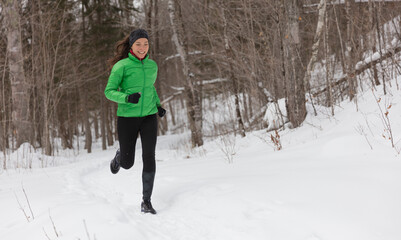 Running in Winter snow. Woman runner jogging outside on cold winter day in forest. Fit healthy lifestyle concept with beautiful young fitness model. Mixed race Asian Caucasian jogging in full length.