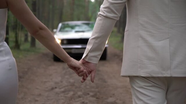 Young couple in love is walking along a forest road. Man and woman hold hands. The bride and groom walk to their car - a white convertible vehicle.