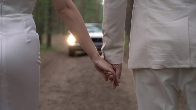 Young couple in love is walking along a forest road. Man and woman hold hands. The bride and groom walk to their car - a white convertible vehicle.