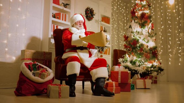 Santa with a real white beard in a traditional red and white suit and round glasses is smiling sitting on a chair and holding a list of children's wishes