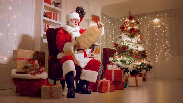 Santa Claus is sitting on a chair reading a list of children's wishes against the background of Christmas lights
