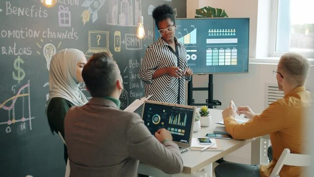Afro-American business lady is making presentation for diverse group of colleagues using electronic board orking in creative office with chalkboard wall. Youth and job concept.