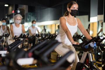 Fototapeta na wymiar Young european female in protective mask riding exercise bike during cycling class in gym